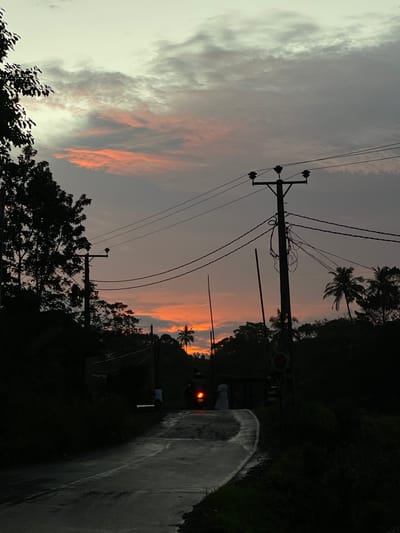 The image depicts a street scene in Kamburugamuwa, Sri Lanka, during dusk or dawn. The sky is visible, showing a mix of gray and orange hues in the clouds. Electrical poles and wires run along the roadside. Vegetation, including palm trees and other foliage, lines the road. A street is visible on the right of the image. In the center of the image, there is a vehicle with its headlights on, and a figure can be seen standing near it. There is no interaction visible between the person and the vehicle. The environment appears to be a rural or semi-rural area.
