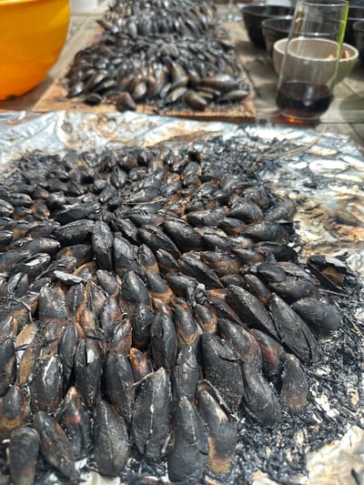 The image shows a close-up of cooked mussels, likely prepared outdoors, resting on a surface covered in aluminum foil. The mussels appear heavily charred, with a dark, almost burnt appearance. A smaller pile of mussels is visible in the background. Some glasses are partially visible in the upper-left corner of the frame. While the location provided is La Couarde-sur-Mer, France, there is no information in the image itself to confirm that this is where the photo was taken.