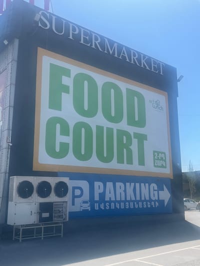 A large, vertically oriented outdoor sign is affixed to a building, indicating various commercial establishments. The sign features multiple sections. On the far left, running vertically, is the word "SUPERMARKET" in white capital letters against a dark background. The central and largest section displays "FOOD COURT" in prominent green capital letters on a white panel with a yellow border. Above "FOOD COURT" are smaller text elements: "artch Lunch" with a logo and "SINCE 2002" in English, and "2-րդ ՀԱՐԿ" (2nd floor) in Armenian. To the right of the food court section is a blue panel with a white arrow pointing right, the word "PARKING" in white capital letters, and the Armenian translation "ԱՎՏՈԿԱՅԱՆԱՏԵՂԻ" (Parking) along with a car icon. Below this parking section, mounted on the building wall, are several industrial air conditioning or refrigeration units, with "LOGIC" and numbers visible on one of them. The sky above is clear and blue, indicating daytime. In the distant background, a white car is visible on what appears to be a paved area or road. The scene suggests a commercial complex in Ayntap, Armenia, offering a supermarket, food court, and designated parking.