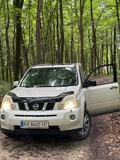 A white Nissan X-Trail SUV, with its driver's side door open, is parked on a dirt path in a forest. The vehicle's headlights are illuminated, and its license plate reads "BX 8662 IC" with a Ukrainian flag emblem. The car is positioned at an angle, facing slightly towards the viewer. The surrounding environment consists of a dense forest with numerous tall trees, their branches forming a canopy overhead. The ground is covered with dirt, fallen leaves, and some scattered stones. The scene appears to be in a natural, wooded area, consistent with a rural location in Kolubaivtsi, Ukraine. There are no people visible in the image.