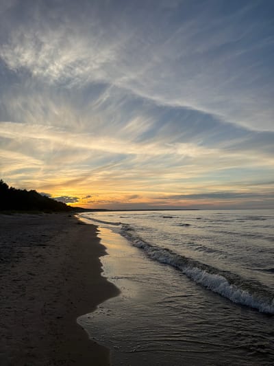 This media file depicts a sunset over the Baltic Sea in Plieņciems, Latvia. The image is oriented vertically, showing the sky and the sea side-by-side. The sky is filled with wispy, elongated clouds that are illuminated by the setting sun, creating a gradient of colors from pale blue to warm yellow and orange near the horizon. A thin line of dark trees forms the silhouette of the coastline. The sea occupies the right side of the frame, with gentle waves rolling onto a sandy beach. The wet sand reflects the colors of the sky, creating a shimmering effect. The overall mood is serene and peaceful, characteristic of a sunset at the beach. There is no visible text in the image.