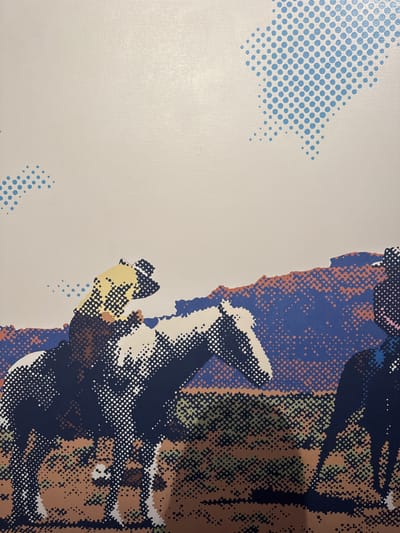 A close-up captures a portion of a graphic print. The image features a section of a halftone-style portrait, with areas of blue, brown, and white dots forming the details of what appears to be a face and clothing. The background is a light neutral color, decorated with scattered patterns of blue dots in varying densities. The style resembles pop art, with its use of bold colors and dot matrix techniques.