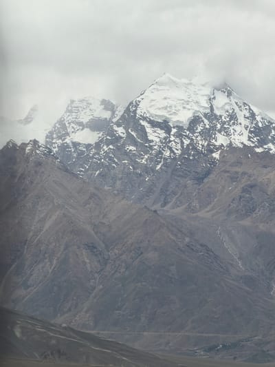 A vast mountain range dominates the frame, with snow-capped peaks piercing through a heavy, overcast sky. The lower slopes of the mountains are a muted gray, textured with rock and scree. A winding, lighter-colored path, possibly a riverbed or road, is visible on the right side of the mountains. In the distance, at the base of the mountains, a cluster of small buildings can be seen, suggesting a settlement. The foreground is a blur of muted, undulating terrain, hinting at foothills or a valley floor. The overall atmosphere is one of rugged, imposing natural beauty, with a stark contrast between the bright, snow-covered summits and the shadowed, rocky lower regions. The image appears to be taken from a distance, capturing the grandeur of the landscape. There are no people or animals visible in the scene. The location is identified as Stongde, India.