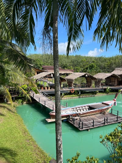 This image captures a serene waterside scene in Tambon Pa Kae Bo Hin, Thailand. The foreground is dominated by lush green palm fronds and a thick palm tree trunk, suggesting a tropical environment. Beyond the foliage, a vibrant turquoise body of water stretches out, with a traditional wooden boat docked alongside. The boat is long and narrow, painted with colorful stripes and featuring wooden decking and railings.
Along the water's edge, a series of rustic structures with thatched roofs are visible, connected by wooden walkways. These buildings appear to be part of a resort or dining area, with tables and chairs set up under open-air roofs. A small orange life preserver is attached to a post near one of the walkways. In the background, a dense line of green trees creates a natural backdrop, with a clear blue sky dotted with a few white clouds overhead.
The lighting suggests it is daytime, likely late morning or early afternoon, with the sun casting shadows and highlighting the textures of the wood and vegetation. The overall atmosphere is tranquil and inviting, evoking a sense of relaxation and tropical escape. There is no visible text in the image.