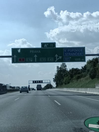 The image shows a highway scene in Všechromy, Czechia, on a partly cloudy day. The view is from a vehicle moving forward on the road. Above the highway, a large green directional sign provides information about routes and destinations.
The sign indicates that "PRAHA" (Prague) is straight ahead, with exits for "E50," "E55," and "E65." To the right, the sign directs towards "V. POPOVICE" and "STRANČICE," with a route number "107" and a distance of "1000 m." Another smaller sign at the top of the larger sign shows the number "15."
Several cars are visible on the highway, including a silver sedan in the foreground and a white van further ahead. The road has clear lane markings. On the right side of the highway, a grassy embankment lined with a concrete barrier is visible, with trees forming a dense line along the top. In the distance, some industrial or construction structures can be seen. The overall impression is of a typical highway commute under a bright sky with scattered clouds.