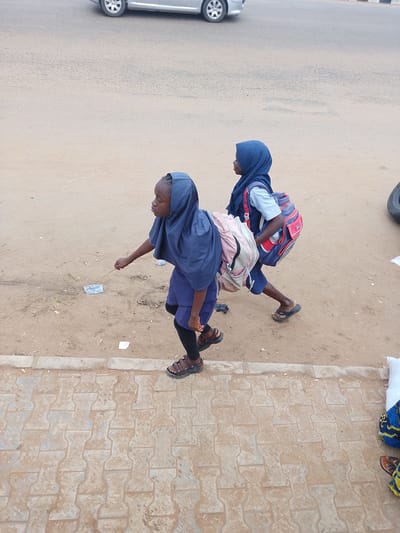Two young girls in blue hijabs are walking on a dirt path next to a paved area. The girl in the foreground is wearing a dark blue uniform and black leggings, with brown sandals. She is holding a stick and appears to be interacting with something on the ground. The girl behind her is wearing a light blue shirt with her blue hijab, a blue skirt, and black leggings, also with brown sandals. She has a red and blue backpack on her back, and a pink and white backpack slung over her shoulder. The ground is a mix of dirt and scattered debris, with a patterned paved walkway in the foreground. In the background, a silver car is visible on a paved road. The scene appears to be outdoors, possibly during the day, with natural lighting. There are no visible signs of text in the image. The overall impression is of children on their way to or from school in a somewhat dusty, urban or semi-urban environment.