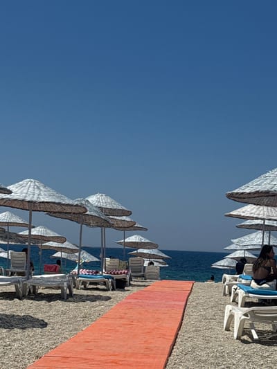 This is a vertical, eye-level shot of a beach in Foça, Turkey, under a clear, bright blue sky. In the foreground, a vibrantly colored orange walkway extends diagonally across the frame from the bottom right to the middle left. The walkway is made of what appears to be wooden planks. To the right of the walkway, several white plastic lounge chairs are visible on a pebbly beach, some with blue cushions. On the left side of the walkway, more lounge chairs are arranged, with some partially obscured by large, conical thatched umbrellas.
In the middle ground, the blue sea stretches towards the horizon. Beyond the beach, there are rows of thatched umbrellas providing shade. A few people are present on the beach, some sitting in lounge chairs or standing. On the far right, a woman with long dark hair is sitting on a lounge chair facing away from the camera, wearing a bikini. Further back, near the water's edge, a person is partially visible. The overall scene depicts a typical beach resort environment on a sunny day.