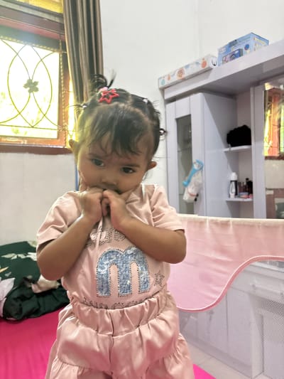 A young girl with dark hair styled in pigtails and a red star clip stands in what appears to be a bedroom in Blangseureukuy, Indonesia. She is wearing a pink t-shirt with a large, sparkling blue letter "M" on it, and matching pink ruffled shorts. Her hands are clasped together near her mouth, and she is looking downwards with a thoughtful expression. The background shows a window with decorative glass panes, a brown curtain, a white cabinet with shelves containing various items, and a pink child's bed or playpen. A green and white patterned blanket is visible on a bed to the left of the frame.