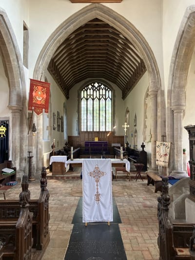 The image displays the interior of St Mary's Church in Mendlesham, Mid Suffolk, United Kingdom, viewed from the nave looking east towards the chancel and altar.
The scene is devoid of people. The church features a high, vaulted wooden beam ceiling and pointed stone arches supported by sturdy pillars on both sides, defining the nave. The floor is constructed of reddish-brown brick tiles, with a dark rectangular stone slab, possibly a tombstone with illegible engraved text, visible centrally in the foreground. Ornate dark wooden pews or choir stalls with carved details line the foreground on either side.
Centrally placed in the midground is a white rectangular banner on a wooden stand, embroidered with a stylized cross in gold and brown tones. Beyond this, the chancel is slightly raised and delineated by a wooden rail. Two smaller altars or tables, draped in white cloths and adorned with candlesticks, flank a main central altar covered with a purple cloth. Behind the main altar, the lower wall is paneled in dark wood.
The eastern wall features a large, multi-paned Gothic-style window with clear glass, through which natural light enters, illuminating the chancel. Trees are visible outside the window.
On the left side of the nave, a red and orange banner hangs from a pillar, displaying the Latin text "O SALUTARIS HOSTIA ADOREMUS IHS" along with religious symbols. Further left, a dark metal gate with a decorative "MR" monogram (likely Marian) is visible. On the right side, another banner is displayed, reading "St Mary's Pilgrim Mendlesham," affirming the church's identity. A plain wooden bench and a dark wooden cabinet-like structure are also present on the right. A tall, free-standing gold cross on a dark pole is positioned to the right of the chancel.