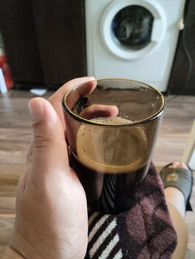 A person's hand is holding a glass of coffee with foam on top. The hand is resting on a maroon and white striped cloth. In the background, a white washing machine is visible. The scene appears to be indoors, possibly in a home. The location in Андрэеўшчынскі сельскі Савет, Belarus, is not evident from the image.