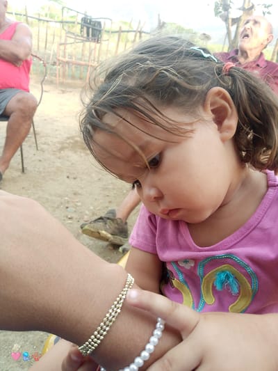 In El Cercado, Venezuela, a close-up shot focuses on a young child wearing a pink shirt with embroidered details. The child's head is tilted downwards, revealing a portion of their face and hair, which is tied back with a red elastic band and a blue clip. The child's left arm, adorned with a gold beaded bracelet, is prominent in the foreground. A second, pearl bracelet is visible on the child's right hand. In the background, an older person in a red tank top and grey shorts is seated, and another elderly person in a purple shirt is visible in the upper right corner of the image, appearing to be speaking. The setting appears to be outdoors, with a dirt ground and a wooden fence in the background, suggesting a rural or semi-rural environment.