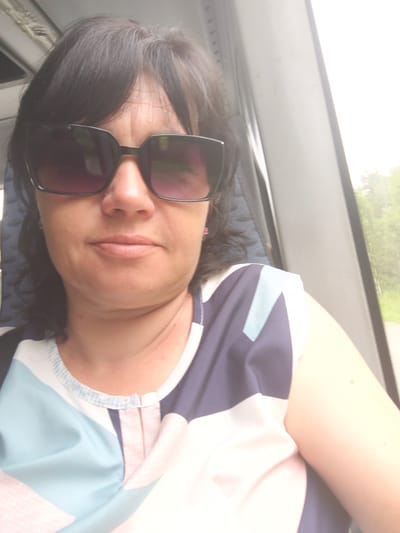 A medium shot photograph shows a middle-aged woman with dark hair and sunglasses, wearing a blue, white, and pink patterned top. She is looking at the camera with a neutral expression. The background is out of focus, but appears to be the interior of a vehicle, possibly a bus, with a window showing greenery outside. The lighting is bright, suggesting it is daytime. There is no specific detail that places this image in Litvinovka, Russia.