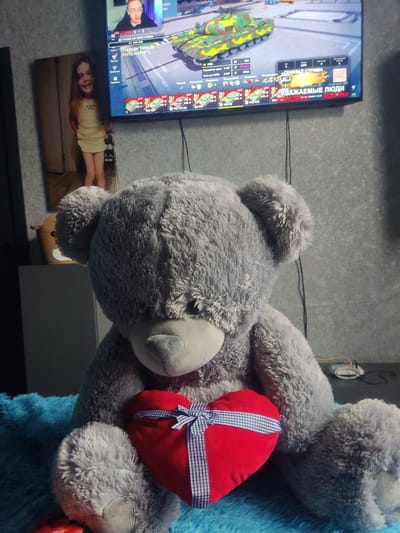 A large, fluffy grey teddy bear is seated, holding a red heart-shaped pillow with a blue and white checkered ribbon tied around it. The bear is placed on a bright blue, fuzzy surface. In the background, a television screen displays a video game interface with tanks and game statistics. To the left of the television, a framed picture of a young girl in a yellow dress is leaning against the wall. The wall appears to be textured. A small woven basket resembling a bear's face is visible on a shelf behind the teddy bear. The overall setting suggests an indoor environment, likely a living room.