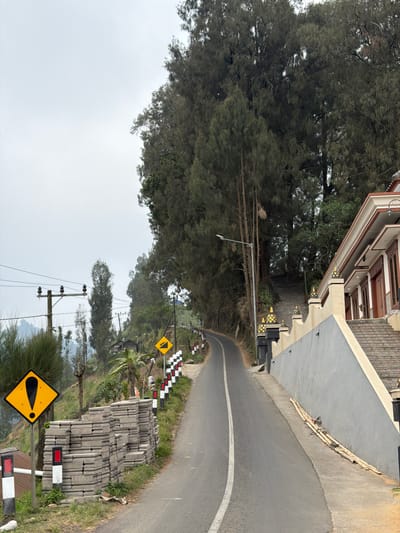 The image depicts a road winding through a hilly, tree-covered landscape in Ngadas, Indonesia. On the right side of the road, a building with traditional Indonesian architecture is visible, featuring a tiled roof and ornate decorative elements. A set of stairs leads up to the building. A low, grey concrete wall runs alongside the road, decorated with black posts topped with yellow finials. Further down the road, a series of red and white striped posts line the side.
To the left of the road, a yellow diamond-shaped sign with a black exclamation mark is prominently displayed. Next to it, stacks of grey, rectangular construction materials are piled neatly. A telephone pole with insulators stands on the left side of the road, with electrical wires extending from it. The road itself is paved and has a white line marking the center. The environment is characterized by lush greenery, with tall, dense trees forming a backdrop. The sky is overcast.