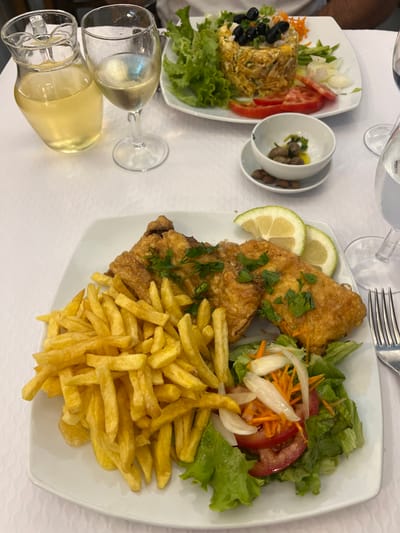 This image depicts a restaurant table in Coruche, Portugal, set for a meal. The table is covered with a white textured tablecloth. In the foreground, a white rectangular plate holds three pieces of golden-brown breaded and fried fish, garnished with fresh parsley. Accompanying the fish is a large portion of golden french fries and a small side salad consisting of green lettuce, sliced red tomatoes, shredded carrots, and sliced white onions. Two lemon slices are placed on the fish.
In the background, another white rectangular plate features a cylindrical serving of a dish, likely a mixed-rice or shredded cod preparation (e.g., *Bacalhau à Brás*), topped with black olives and green garnish, surrounded by lettuce, sliced tomatoes, and onions. Between the plates, a small white bowl on a saucer contains green olives in olive oil, with several brown-shelled almonds placed beside it.
To the left, a clear glass pitcher holds a pale yellow liquid, identified as a beverage such as white wine or sangria, next to a wine glass containing the same liquid. A second wine glass, partially visible on the right, appears to hold a clear liquid. A silver fork is visible to the right of the foreground plate. A person's arm is partially visible in the upper right background, indicating a diner.