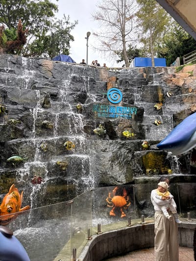 This image displays an artificial rock face waterfall at the Qingdao Underwater World in Shinan District, China, under an overcast sky. The dark grey, multi-tiered rock face features numerous cascading water flows and is adorned with various stylized, colorful marine life sculptures, including fish and crabs. Centered on the rock face is a blue spiral logo positioned above the Chinese characters "青岛海底世界" and the English text "Underwater World."
In the lower right foreground, an adult wearing a light-colored top and wide-leg beige pants holds an infant, who is dressed in a light hat and yellow bib. They are standing behind a clear glass barrier with silver posts, which separates the viewing area from the waterfall's base. Partial views of large blue and white marine animal statues, specifically a dolphin's head on the far left and a shark's head on the far right, extend into the scene. A prominent large orange fish sculpture is visible near the lower left.
On a platform situated above the waterfall, approximately five individuals are visible standing behind a railing. This upper level is bordered by green foliage and several bare deciduous trees. A streetlamp is present on the platform, and a blue tarp or tent-like structure is partially visible on the far left of this upper tier.