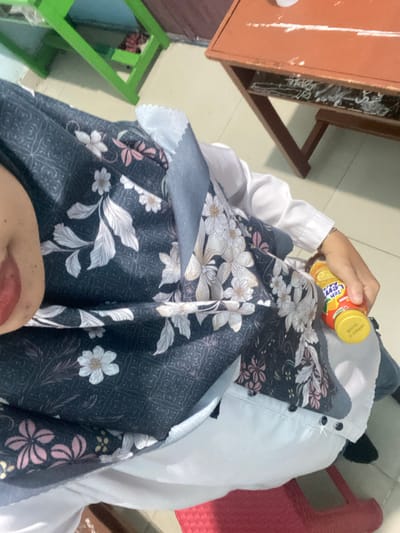 The image shows a person holding a small bottle of "Teh Lemon" drink while sitting in front of a wooden table. The person is wearing a white shirt and a dark patterned hijab with floral designs. The background includes a green shelf and a floor. The scene appears to be indoors, possibly in a classroom or study area. The context of Tanjung Gading, Indonesia, is not directly discernible from the visual elements of the image.