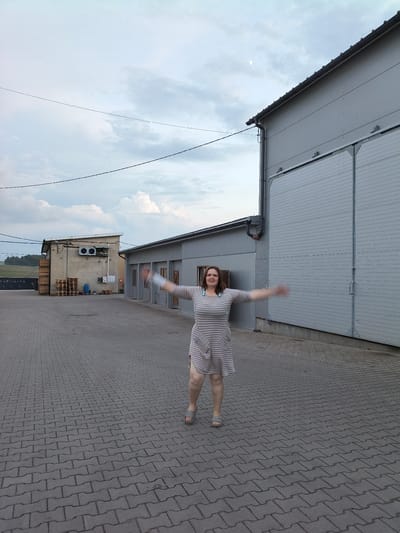 In the city of Galonki, Poland, on a cloudy day, a woman stands in a paved courtyard, her arms outstretched. She is wearing a striped dress and sandals. In the background, there are two industrial buildings, one with large metal doors and the other with air conditioning units and a stack of wooden pallets. Electrical wires are visible against the sky, and a crescent moon hangs in the upper left corner of the image. The ground is covered with interlocking gray paving stones.