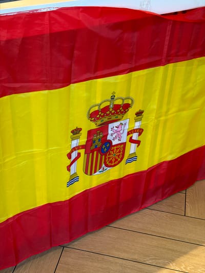 The image shows a close-up view of the Spanish flag draped over a surface, likely a table or counter. The flag is predominantly red and yellow, with the coat of arms of Spain displayed in the center. The coat of arms features a shield divided into quadrants, a crown, and pillars. The flag appears to be made of synthetic fabric and is slightly creased, indicating it is not perfectly flat. The background is out of focus, but a glimpse of wooden flooring is visible at the bottom right of the image. There are no people or animals present in the image. The lighting suggests an indoor setting. There is no information in the image to indicate the location is in Húmera-somosaguas-prado Del Rey, Spain.