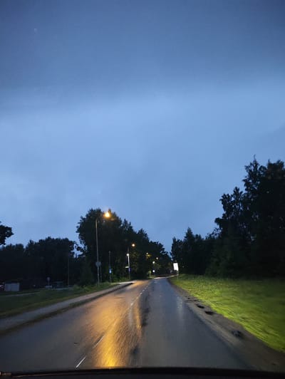 The image depicts a scene on a wet road in Harkujärve, Estonia, during dusk or pre-dawn. The road surface is reflective, showing streaks of light from streetlights and possibly headlights. Trees line both sides of the road, with some appearing as dark silhouettes against the muted sky. Streetlights are visible along the road, casting a warm, yellowish glow. The sky is a deep blue-grey, suggesting overcast conditions or the transition between day and night. There are no people or animals visible in the frame. The overall impression is one of a quiet, damp evening on a suburban road.