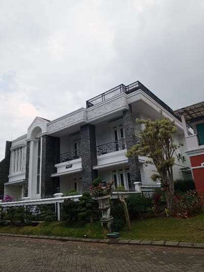 This image displays a multi-story residential building located in Weintiapa, Liberia, under an overcast sky. The structure features a predominantly white facade accented with dark grey stone cladding on pillars and sections. Architectural details include an arched entrance with fluted pilasters on the left, decorative friezes, and white-framed windows, some of which are balcony access doors. Multiple balconies with black wrought-iron railings are visible on the second floor, one bearing a "NO 1109" plaque. A rooftop terrace, also secured with black wrought-iron railings, is present on the uppermost level. Several exterior air conditioning units are mounted on the right side of the building. The property is demarcated by a white picket fence, behind which lies a manicured lawn, various shrubs, and a prominent deciduous tree with light green foliage. A decorative concrete urn containing red and green plants stands on the lawn in the mid-foreground. The immediate foreground consists of an interlocking concrete paver driveway or street, bordered by a concrete curb. No individuals or dynamic actions are present in the scene.