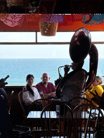 In Puerto Nuevo, Mexico, patrons are seated at tables overlooking the ocean, with a tuba player performing in the foreground. The scene is dimly lit with overhead lighting and natural light filtering through a large window. The tuba player, wearing a plaid shirt, is silhouetted against the bright ocean view. A woman in a white shirt and a man in a maroon shirt are seated at a table in the middle ground, facing the camera. A woman in a yellow shirt is visible to the right. Festive paper decorations hang overhead. Wooden chairs and tables are arranged in the seating area. The overall atmosphere appears to be a relaxed, possibly musical, dining experience by the sea.