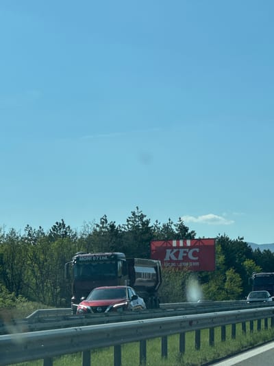 The image depicts a daytime scene on a multi-lane highway, observed from a vehicle. The sky is clear and bright blue with a single small white cloud.

To the right of the highway, a large red billboard is prominently displayed, featuring the "KFC" logo in white. Below the logo, smaller white text reads "KFC.BG" and a phone number, "8709 11 999". A white glare or lens flare is visible on the billboard's surface.

Vehicles are traveling on the highway. A large dark truck, with "MONI 07 Ltd." visible on its side, is in the foreground. Behind the truck, a red SUV, possibly a Nissan model due to its headlight design, is also present. Other vehicles are visible further down the road, indicating traffic.

Along the right side of the highway, dense green trees and vegetation line the route, with mountains or hills visible in the distant background. A metal guardrail separates the highway lanes from the roadside. The setting appears to be a sunny day in Lesichovo, Bulgaria, consistent with the "KFC.BG" on the billboard.