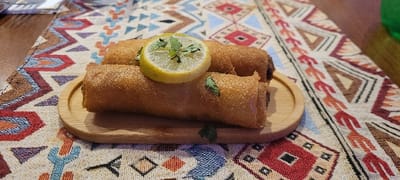 Two golden-brown spring rolls lie atop a small, round wooden tray. A slice of lemon, garnished with sprigs of fresh herbs, sits atop the rolls. The tray rests on a patterned textile featuring geometric shapes in earth tones like brown, red, and blue, with a wooden table visible in the background. The lighting highlights the crispy texture of the rolls.