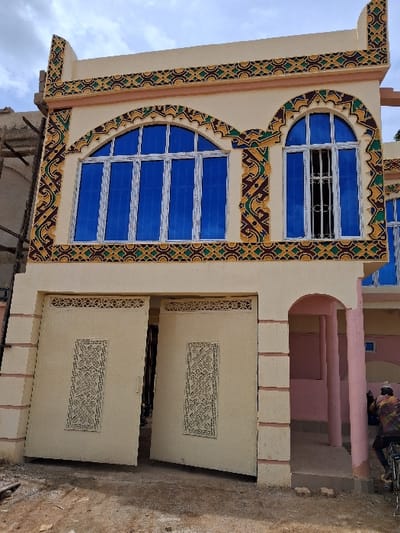 A two-story building in Madobi, Nigeria, features a cream-colored facade with intricate geometric patterns in yellow, green, brown, and black around its upper windows and roofline. The upper level contains two arched windows with blue-tinted glass and white frames; the larger left window has five panes, while the right window has two panes and is fitted with a white metal security grille. The ground level includes a large, light-colored double metal gate, partially ajar, revealing a dark interior beyond. The gate panels are embellished with cut-out scrollwork designs. To the right of the gate, an arched entryway supported by pink columns is visible. A person on a bicycle, clad in a patterned multi-color shirt, is partially visible on the far right, positioned near the building's base. The foreground consists of unpaved dirt ground. The sky above is partly cloudy. A scaffolding-like structure is partially visible on the left side of the building.