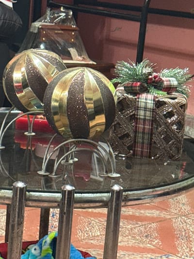 The image shows a still life on a glass-topped metal table. Two brown and gold Christmas ornaments rest on the table, near some red candles. A Christmas decorative item with greenery and a plaid ribbon is positioned near the center of the table. A wrapped wooden box is partly visible behind the ornaments. The background features a pinkish-red wall and other furniture. There is no indication as to the image's connection to San Cristóbal, Mexico. No people are visible.