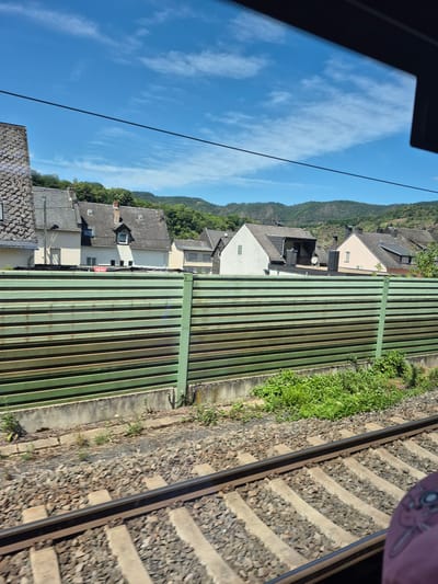 This image depicts a view from a moving train through a window, showcasing a town and surrounding landscape in Kobern-Gondorf, Germany. In the foreground, railway tracks with concrete sleepers and ballast are visible, suggesting the train's forward motion. A green, horizontal-slatted fence or barrier runs across the middle ground, partially obscuring the view of houses and hills. Behind the fence, a row of residential buildings with tiled roofs, chimneys, and windows can be seen. The houses are of varying sizes and architectural styles, with some appearing older and others more modern. Beyond the houses, lush green hills covered in trees form the backdrop. The sky is a clear, bright blue with scattered white clouds. The overall scene suggests a sunny day in a rural or semi-rural setting.