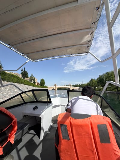 A bright, sunny image captures a scene aboard a small boat cruising a waterway in Kuchtumgali, Uzbekistan. The perspective is from behind a seated individual, likely operating the boat, who wears a white short-sleeved shirt and a prominent orange life vest with two black rectangular patches on the back. The boat's structure includes white metal railings and a light-colored canvas canopy above, secured by grommets and ropes.
The surroundings reveal a serene landscape under a clear blue sky dotted with scattered white clouds. On the right, the waterway is bordered by a well-kept embankment lined with lush green trees and what appears to be a stone or paved path. In the distance, architectural structures, possibly part of a bridge or buildings, are visible along the bank. The water surface is calm, reflecting parts of the sky and trees.
The scene conveys a peaceful journey during daylight hours, characterized by clear weather. No discernible text is visible in the image.