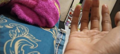 This media file shows a close-up, first-person perspective of a hand reaching towards the camera. The hand is lightly tanned and appears to be resting on or near a surface covered with a blue fabric featuring a white ornate pattern. To the left of the hand, a folded pink fuzzy blanket is visible. In the background, out of focus, there's a shelving unit with several items, possibly bottles or containers, suggesting an indoor setting. The lighting appears to be artificial, casting a warm glow. There is no visible text in the image. The overall scene suggests a casual, everyday moment. The location context provided is Duman, Indonesia.