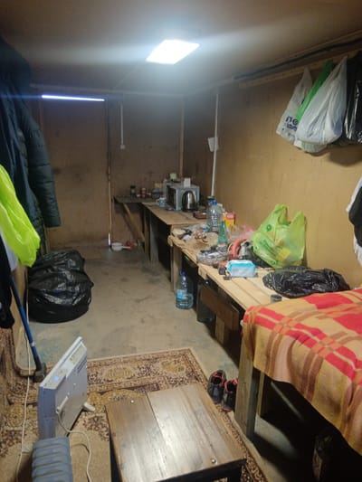 The image shows the interior of a room located in Яльгелево, Russia. The room appears cluttered and sparsely furnished.
**Environment:** The walls are made of unpainted wood. A single square light fixture is mounted in the ceiling. The floor is partially covered by an oriental-style rug.
**Objects:** A wooden table occupies the center of the room. An electric radiator stands near the table. Along one wall, there is a countertop cluttered with various items, including a microwave, kettle, food containers, tools, and bottles. A bed with a red and brown blanket is positioned along another wall. Several plastic shopping bags hang from the ceiling. Bags of garbage are visible in the corner of the room. A pair of running shoes are next to the bed.
**People:** No people are visible in the image.
**Actions/Interactions:** None observed.