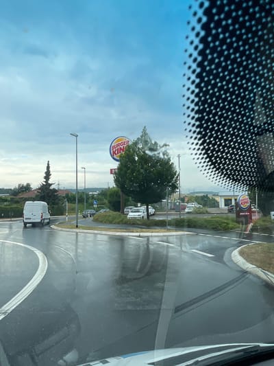 A wet, overcast day is depicted, possibly in Lauterbach, Germany, with the Burger King logo prominently visible on a large sign in the mid-ground. The foreground shows a wet road with white lane markings, suggesting a roundabout or intersection. A white van is turning in the roundabout. A Burger King restaurant is visible in the background, with a drive-thru sign. Trees and buildings are scattered throughout the landscape, with power lines and hills in the distance. The image is taken from inside a vehicle, with a dotted pattern from the windshield obscuring the upper right portion of the frame.