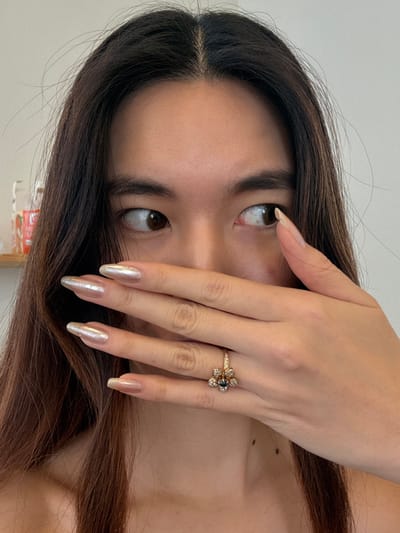 The image captures a close-up, chest-up view of a female individual located in an indoor setting within Arcueil, France. The subject, of East Asian descent, possesses long, dark brown hair with lighter undertones, parted centrally. Her brown eyes are visibly directed to the viewer's right. Her right hand is positioned horizontally across her lower face, covering her mouth and nose. The fingernails on her right hand are long, almond-shaped, and coated with a shimmering, iridescent light peach-gold polish. A gold-toned ring, featuring a central dark stone surrounded by smaller clear stones arranged in a floral pattern, is worn on the ring finger of her right hand. The subject's bare left shoulder is partially visible. The background consists primarily of a plain white wall, with indistinct, out-of-focus objects visible on the far left edge.