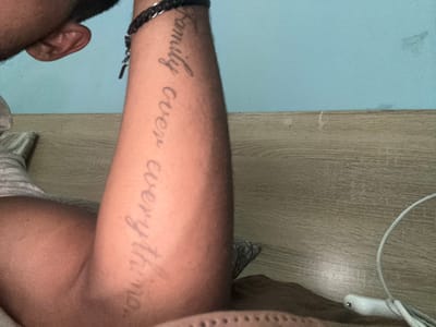 This is a media file of a person's arm with a tattoo on it. The tattoo says "family over everything". The arm is light-skinned with some hair. The person is wearing a black bracelet around their wrist. The background is a wooden surface and a light blue wall. The image is taken from a close-up perspective, focusing on the arm and tattoo. There are no notable details about time of day, weather, or specific emotions. The overall scene is simple and straightforward, showcasing the tattoo. The location context provided indicates Zlataritsa, Bulgaria, but there are no visual cues within the image itself to confirm this.
