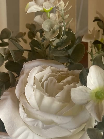 The image shows a close-up of artificial flowers, likely a floral arrangement. The dominant element is a large, creamy white rose with layered petals. To the right of the rose is a smaller white flower with a green center. Behind and around the roses are artificial eucalyptus leaves in various shades of green and gray-green. The background is out of focus but appears to be a vertical striped pattern in muted tones of beige and brown. There is no indication of the city of Важное, Russia, or any specific urban environment. The lighting is soft and diffused.