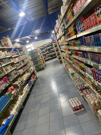 The image presents an overhead, tilted view looking down a long aisle of a well-stocked retail store, likely a supermarket or large grocery shop, located in Goderich, Sierra Leone. No people are visible, and no active event is taking place, suggesting the store is either closed, sparsely populated, or the shot captures an empty moment.
The upper section of the image displays multiple rows of shelves packed with various canned and bottled beverages, including soft drinks, energy drinks, and possibly juices. Brands like "Malta Guinness" are faintly discernible on some cans, a common beverage in West Africa. Further down the aisle, several multi-packs of red-labeled cans, resembling Coca-Cola, are stacked on the blue-tiled floor.
The lower section of the image features a long aisle with open refrigeration or freezer units, likely holding chilled or frozen goods. Beyond these units, more shelves are visible, stocked with a variety of packaged items, which could include dry goods such as rice, pasta, or snacks.
The store's interior features a blue-painted industrial-style ceiling with exposed support beams and bright strip lighting fixtures, some of which create lens flares. The floor throughout the visible area is covered with light blue tiles. A large, rectangular window can be seen in the distant background, providing some natural light and suggesting it is daytime. The overall impression is one of an organized and well-maintained commercial establishment. No specific weather conditions are discernible from the indoor setting.