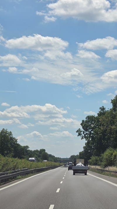 This media file shows a view from a vehicle driving on a highway in Germany, likely near Calau. The perspective is from the passenger side, looking forward and slightly upwards. The scene is dominated by a bright blue sky filled with fluffy white clouds. Lush green trees line the right side of the highway, creating a natural barrier. The highway itself is asphalt with white dashed lines marking the lanes.
Several vehicles are visible on the road ahead, suggesting moderate traffic. The closest vehicle is a dark-colored pickup truck with a white tarp covering its cargo bed. Behind it, other cars and trucks are in motion. The time of day appears to be daytime, with bright sunlight casting shadows and illuminating the scene. The weather seems pleasant, with clear skies and scattered clouds.
The overall impression is a typical highway drive through a scenic, green landscape. There is no visible text in the image. The composition emphasizes the open road and the expansive sky, conveying a sense of travel and freedom. The presence of trees suggests a rural or suburban setting.