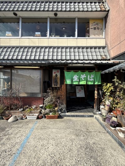 A traditional Japanese building with a gray tiled roof and several windows is visible. Green banners with Japanese writing hang above the entrance, which is decorated with plants in pots. A sign with additional Japanese writing hangs below the banners. The building is adjacent to a paved area with a blue line, likely a sidewalk or street.