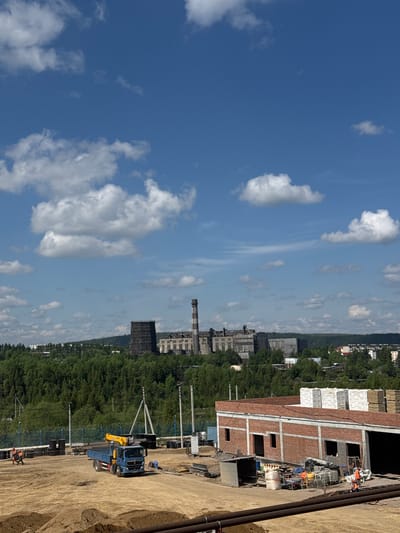 This is an eye-level, full shot of an industrial construction site in Chulman, Russia, under a bright, partly cloudy sky. In the foreground, a blue flatbed truck with a yellow crane is parked on a dirt lot, facing right. Two construction workers in orange vests are visible on the left side of the frame, walking near the truck. A brick building with open doorways and windows is under construction on the right side, with stacks of building materials like pallets of bricks and lumber visible on its roof and beside it. In the midground, a chain-link fence separates the construction site from a line of trees. In the background, a large, sprawling industrial complex with a tall, smokestack-like tower dominates the horizon, nestled among green trees and scattered buildings. The overall impression is one of ongoing industrial development in a forested area.