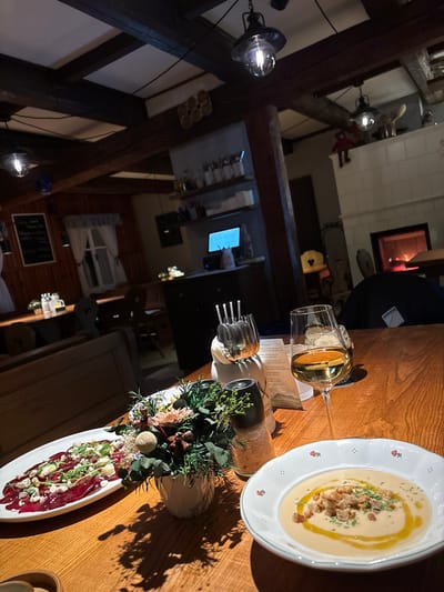 A restaurant table showcases culinary artistry: a plate of creamy soup garnished with herbs and croutons, a delicate carpaccio with greens and cheese, a vase of floral arrangements, and sparkling glassware. Silverware rests neatly beside a menu. The interior features rustic wooden beams, warm lighting, and a glimpse of a fireplace in the background, creating an inviting ambiance.