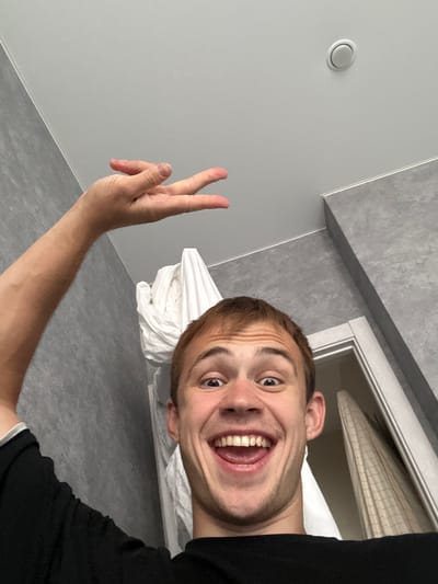 This is a selfie taken by a young man with fair skin and short brown hair. He is wearing a black t-shirt. He is smiling broadly with his mouth open, showing his teeth. His right hand is raised, making a peace sign with his index and middle fingers extended. The background appears to be a bathroom or a room with light grey tiled walls and a light grey floor. A white towel or sheet is visible behind him, and a shower curtain can be seen in the upper right corner. The lighting suggests it is daytime. The image is taken from a low angle, looking up at the man. There is no visible text in the image.