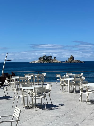 An outdoor seating area of a cafe or restaurant faces the Adriatic Sea in Brežine, Montenegro. White metal tables and chairs are arranged on a light-colored concrete patio. In the distance, a small rocky island dotted with trees and topped with a stone structure is visible in the deep blue sea under a bright blue sky with scattered clouds. A metal railing separates the patio from the water. A portion of a tall, thin white pole with a dark object attached is visible on the left side of the frame. The sun casts shadows from the tables and chairs onto the patio. No people are present in the scene.