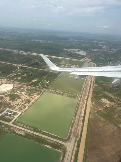 An aerial view captures a landscape with agricultural fields, water retention ponds, and a river, all seen from an airplane. The plane's wing is prominently featured in the foreground, extending from the left side of the image. Below the wing, the earth unfolds with a mix of green vegetation and brown, cultivated land, divided into rectangular plots.
A muddy, brown river or canal runs horizontally across the middle of the frame, bordered by a strip of darker vegetation and a road. Adjacent to this waterway are several large, rectangular ponds filled with murky green water. Some of these ponds have a lighter, sandy perimeter. Further out, the landscape transitions into more densely vegetated areas and what appear to be scattered settlements.
In the distance, a hazy, light-colored sky suggests either a bright day with atmospheric haze or a possibly overcast condition. The overall scene appears to be a rural or semi-rural area, likely agricultural land undergoing cultivation or water management. There is no visible text in the image. The perspective suggests the photograph was taken during daylight hours.