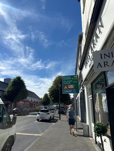 This is a vertical, eye-level photograph taken on a sunny day in Westport, Ireland. The image captures a street scene with a clear blue sky featuring wispy white clouds. In the foreground, a person is walking on a paved sidewalk. A white car is parked in a bay to the right of the road.
The street is lined with buildings, suggesting a commercial or town center. Signs are visible on the buildings, including one that reads "SuperValu" and another with "ROGER & IRONM". A prominent green road sign is hanging over the sidewalk, indicating directions to "Baingear BANGOR N59" and "ath Cliath DUBLIN (N5)". The sign also features symbols for a caravan park and a camping site.
Other cars are parked along the street, and trees with lush green foliage are present, adding to the pleasant atmosphere. The overall impression is of a typical day in a European town, with people going about their activities under a bright sky. The presence of the road sign with Irish place names strongly suggests the location is Ireland, and specifically Westport, as indicated.