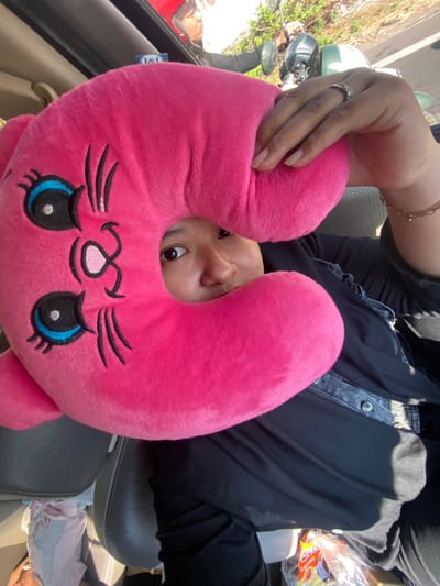 In Bangsal, Indonesia, a person wearing a black hijab and clothing is inside a vehicle, holding up a bright pink neck pillow shaped like a cat's face. The pillow covers most of their face, with one eye peeking through a circular cutout. The person is wearing a ring on their left hand, which is adorned with small stones. They are also wearing a bracelet. In the background, there is another person wearing a red helmet and a green motorcycle. The vehicle appears to be stationary, with the rear of a seat visible in the foreground. Sunlight streams in from the right side of the frame.