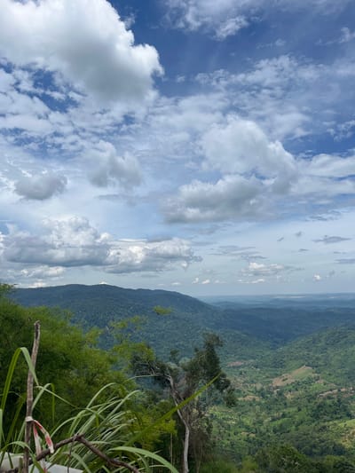 The image depicts a panoramic view of a lush, mountainous landscape. In the foreground, tall green grasses and bamboo stalks are visible, with a weathered wooden post tied with red and white tape. Beyond the foreground, rolling hills covered in dense green foliage dominate the scene, with a deep valley carved between them. In the distance, layers of mountains fade into a hazy horizon under a dynamic sky filled with white and gray clouds against a bright blue backdrop. This vista is characteristic of the natural beauty found in the rural areas surrounding Son Dien, Vietnam, likely from a high vantage point offering a sweeping view of the countryside.
