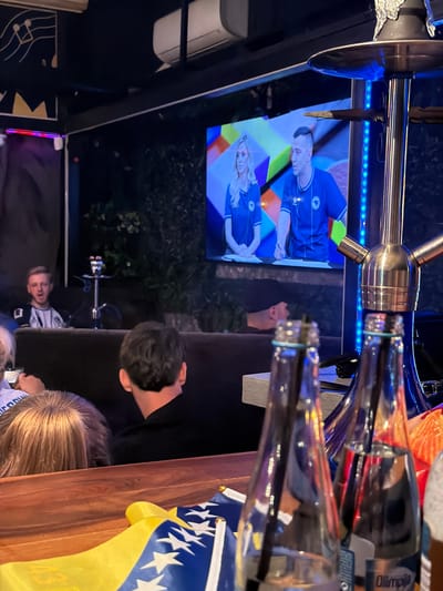 The image depicts an indoor scene, likely a lounge or cafe, with a shisha pipe prominently featured in the foreground. Two glass bottles with blue caps are also visible on a wooden surface. In the background, a large screen displays two individuals, a woman and a man, wearing blue sportswear. Another man with blond hair and a white shirt is seated to the left of the screen, with a shisha pipe behind him. The environment appears dimly lit, with some artificial lighting creating reflections on the glass bottles and the shisha pipe. A flag, partially visible, with blue and yellow colors and white stars, is placed on the wooden surface, suggesting a possible connection to Bosnia and Herzegovina. The scene does not provide specific clues to pinpoint the exact location within the city of Travnik, Bosnia and Herzegovina, beyond the presence of the flag which may allude to national affiliation.
